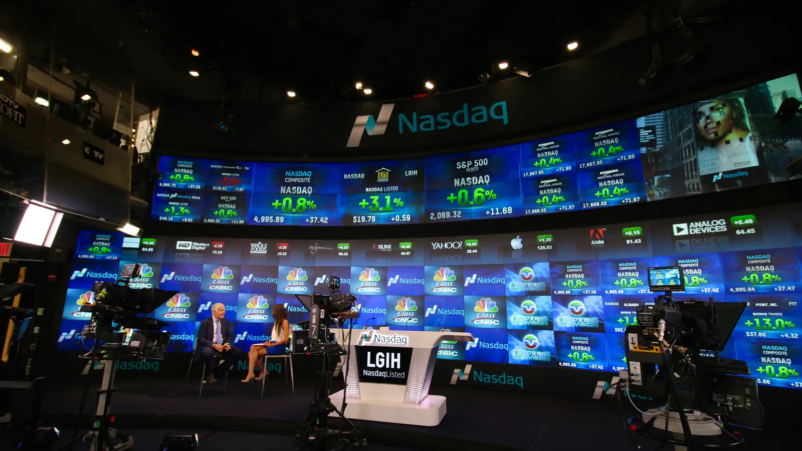 Estúdio do Nasdaq em Nova Iorque com uma parede curva de ecrãs a mostrar cotações e índices, câmaras de televisão em primeiro plano e duas pessoas sentadas numa entrevista