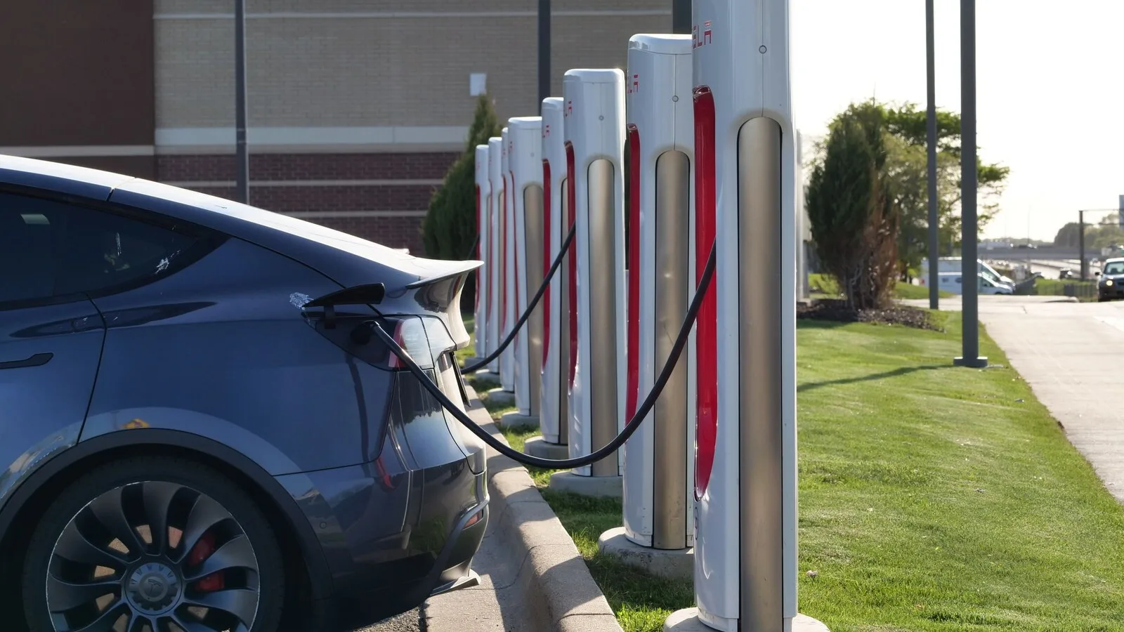 Carro elétrico cinzento ligado a um posto de carregamento rápido, ao lado de uma fila de carregadores brancos e vermelhos junto a um relvado