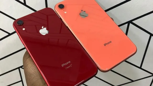 Mão a segurar dois iPhone XR vistos de costas, um vermelho e outro cor de coral, à frente de uma parede branca com linhas pretas