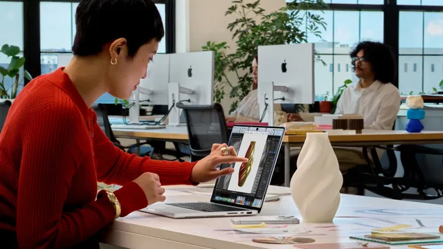 Mulher sentada a uma secretária toca com o dedo no ecrã de um MacBook Pro prateado que mostra um modelo 3D de um vaso