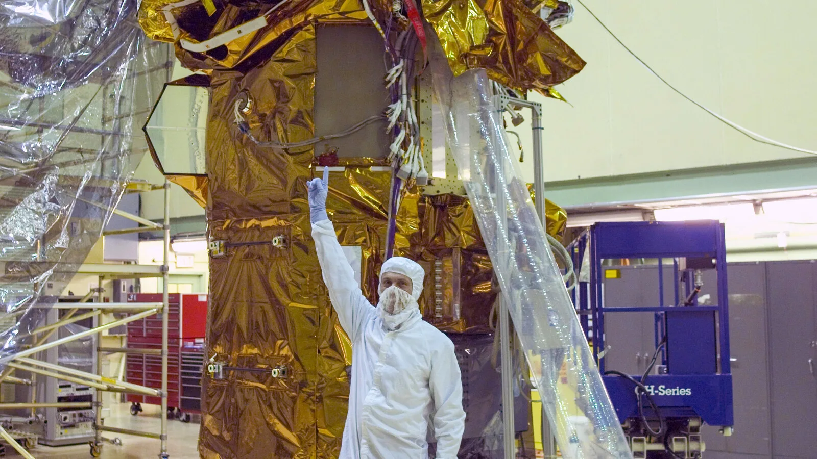 Técnico de fato branco de sala limpa aponta para cima, junto a um satélite coberto de isolamento dourado num pavilhão industrial