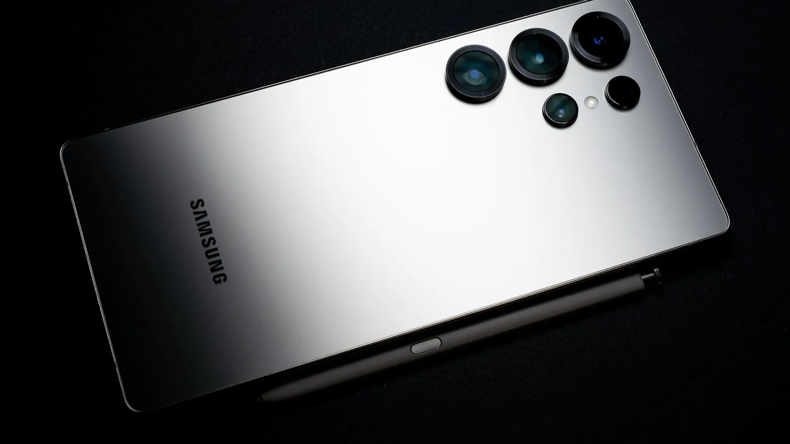 Traseira prateada de um telemóvel Samsung com cinco lentes de câmara, pousado sobre um fundo preto ao lado de uma caneta
