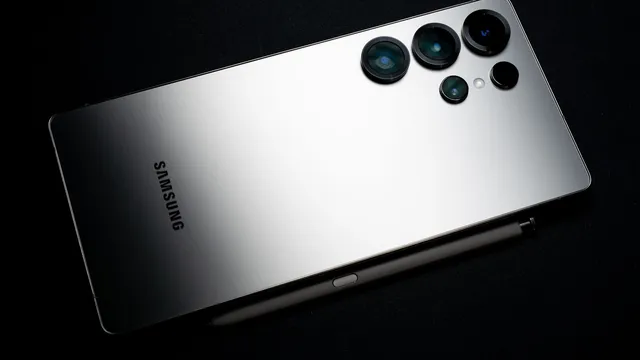 Traseira prateada de um telemóvel Samsung com cinco lentes de câmara, pousado sobre um fundo preto ao lado de uma caneta