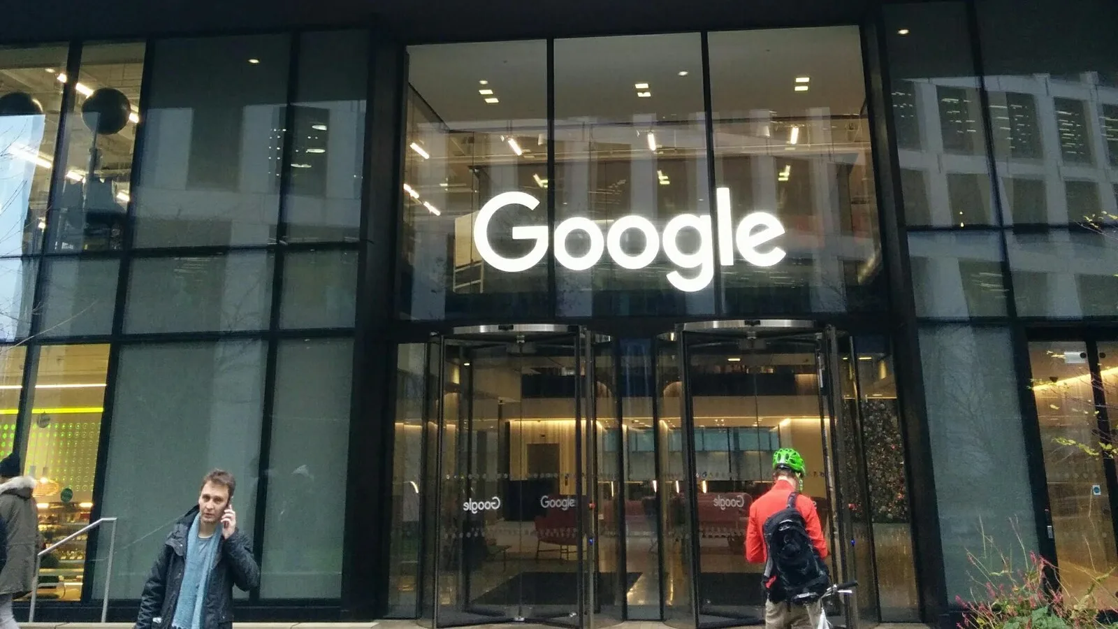 Entrada envidraçada de um edifício com o nome Google iluminado por cima das portas giratórias e duas pessoas no passeio