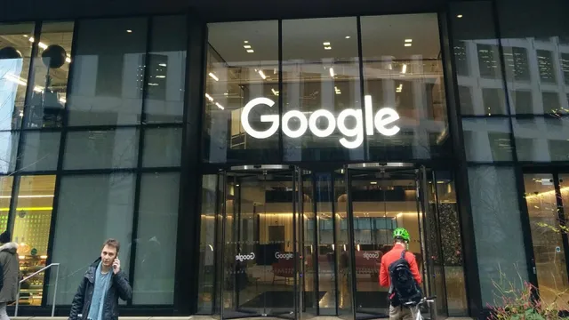Entrada envidraçada de um edifício com o nome Google iluminado por cima das portas giratórias e duas pessoas no passeio