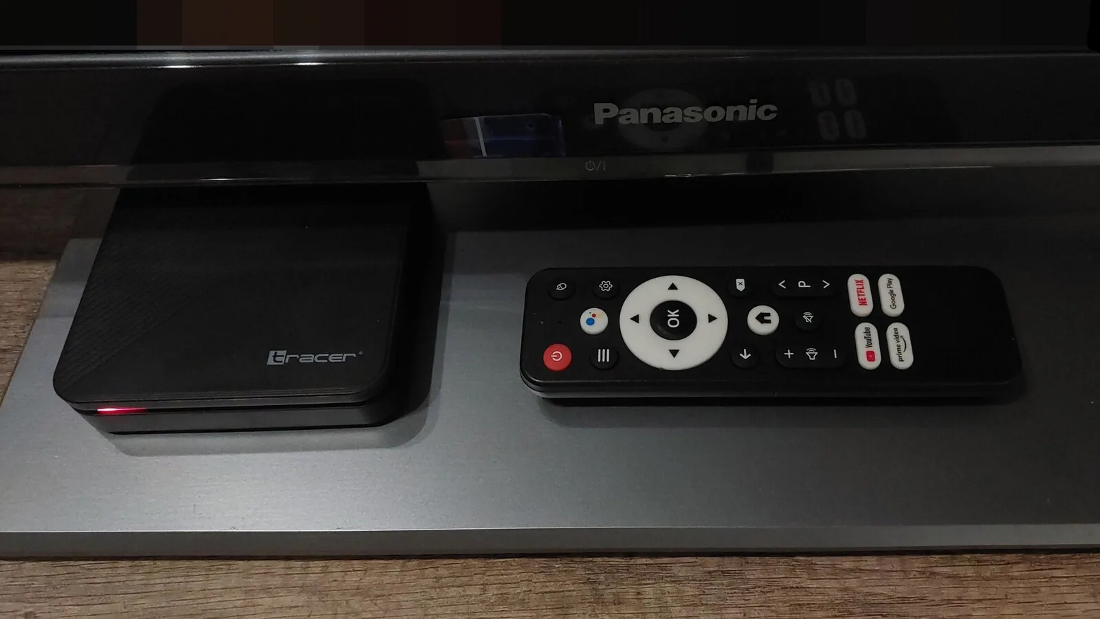 Box Android TV preta pousada num móvel, debaixo de um televisor, ao lado do respetivo comando com botões de serviços de streaming