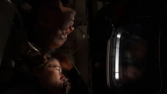 Dois astronautas, um por cima do outro, olham por uma janela da cápsula Orion, iluminados pela luz que entra
