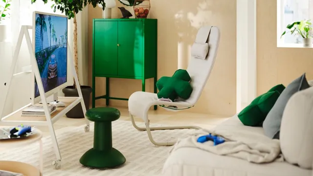 Sala com um móvel de TV branco com rodas, uma poltrona bege com uma almofada verde em forma de cruz, um banco verde em forma de joystick e comandos de jogo pousados no sofá