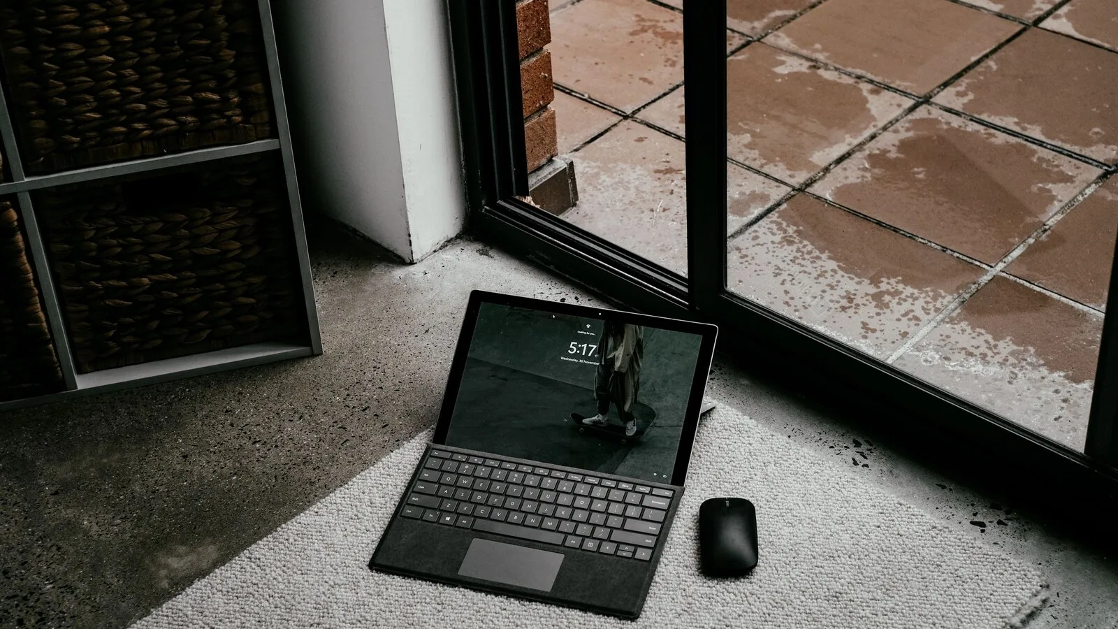 Computador portátil com teclado destacável pousado num tapete junto a uma porta de vidro, com o ecrã de bloqueio ligado e um rato ao lado
