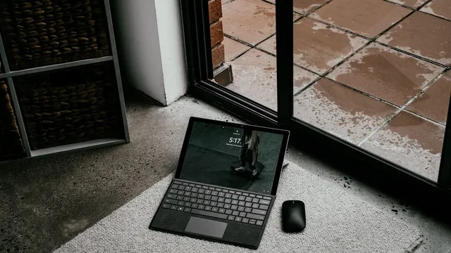 Computador portátil com teclado destacável pousado num tapete junto a uma porta de vidro, com o ecrã de bloqueio ligado e um rato ao lado