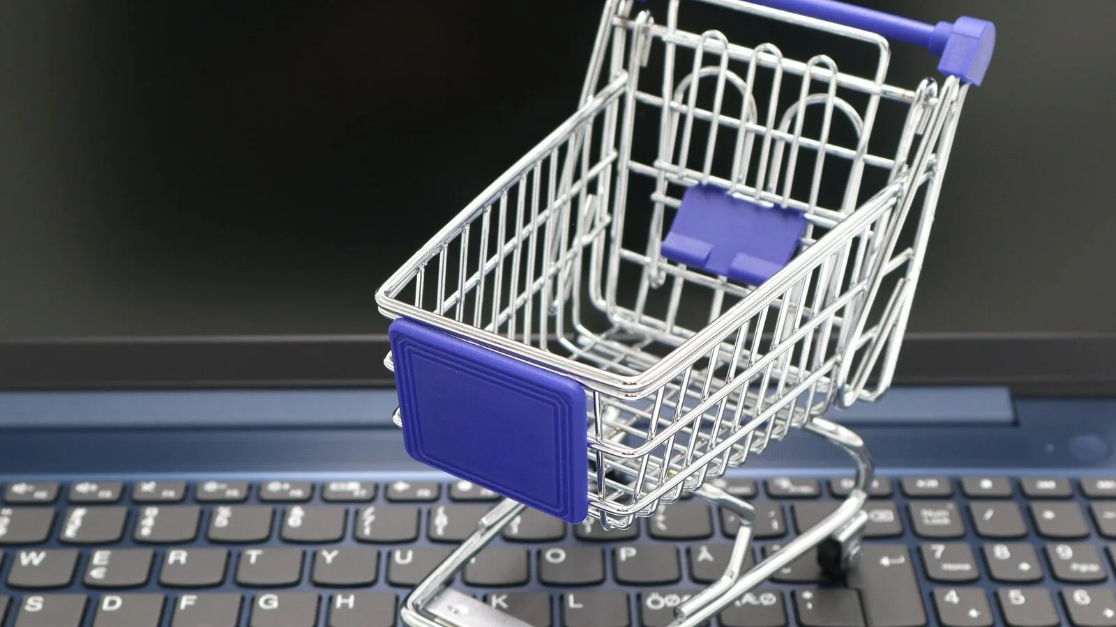 Carrinho de compras em miniatura, metálico e com detalhes azuis, pousado sobre o teclado de um computador portátil