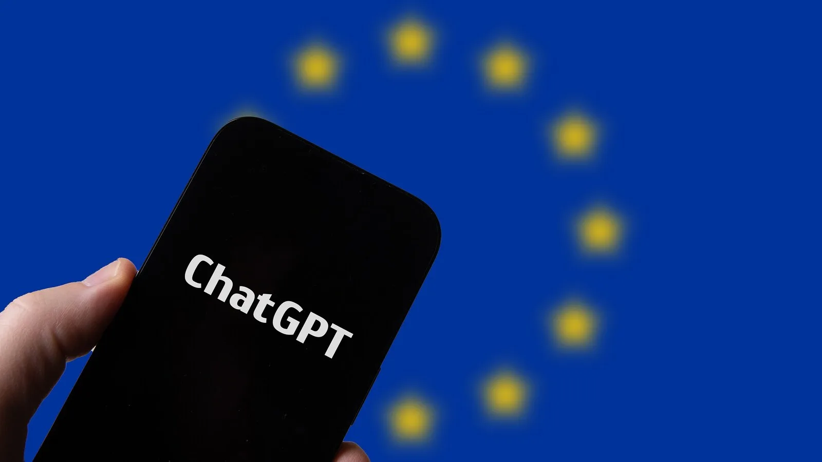 Mão a segurar um telemóvel com a palavra ChatGPT no ecrã, à frente de uma bandeira da União Europeia desfocada