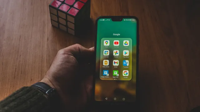 Mão a segurar um telemóvel Android com uma pasta de aplicações da Google aberta, onde se vê o ícone da Play Store, ao lado de um cubo de Rubik