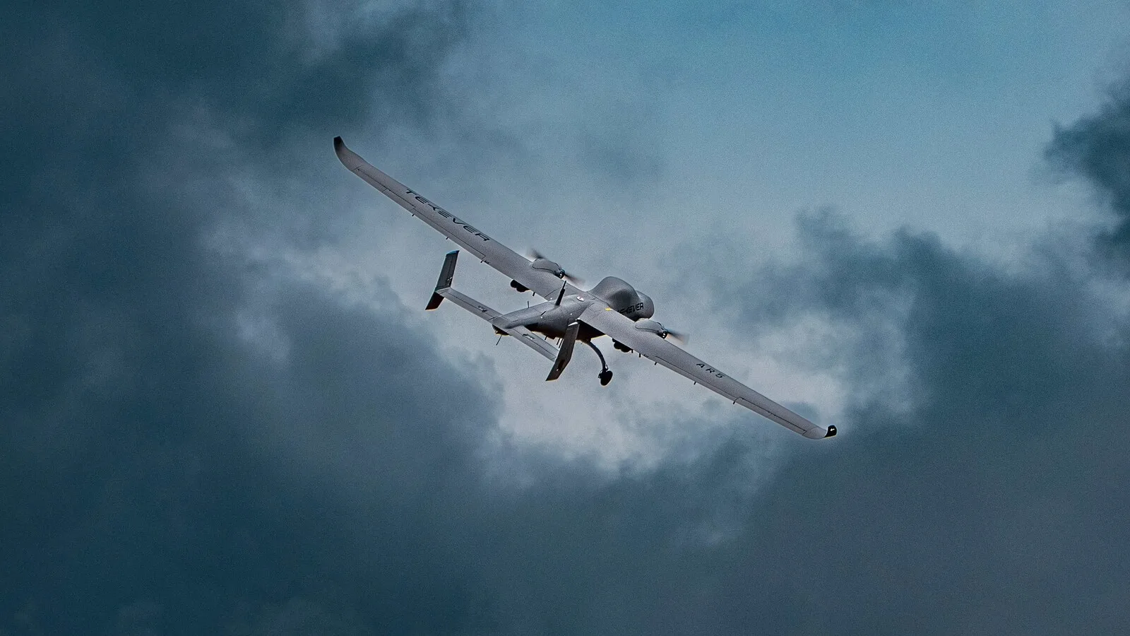 Drone de asa fixa TEKEVER AR5 em voo, visto de baixo, contra um céu carregado de nuvens