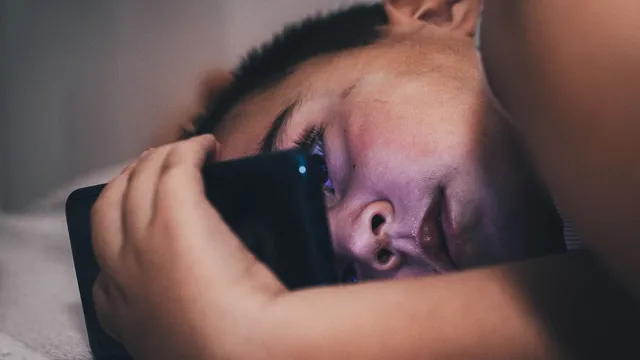 Rapaz deitado na cama, de noite, com o rosto iluminado pelo ecrã do telemóvel que segura junto à cara