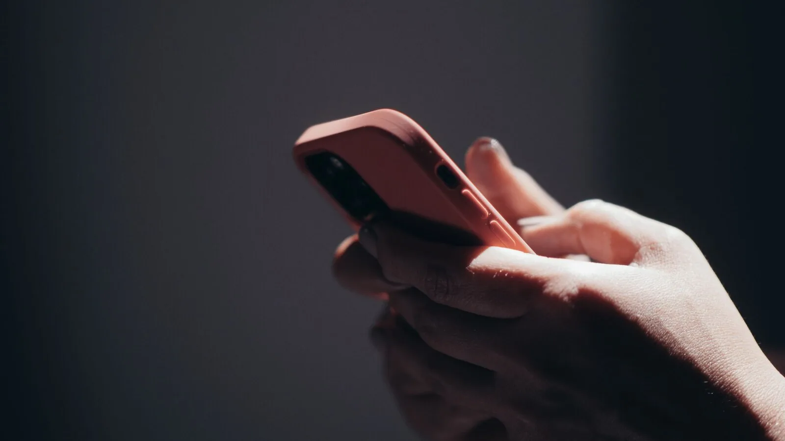 Mão a segurar um telemóvel com capa cor-de-rosa, sobre um fundo escuro