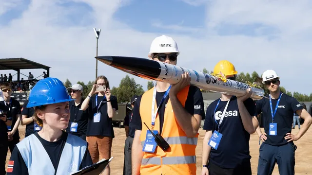 Estudantes com capacetes de obra e coletes refletores transportam ao ombro um foguete branco e preto num terreno de terra batida, com bancadas ao fundo