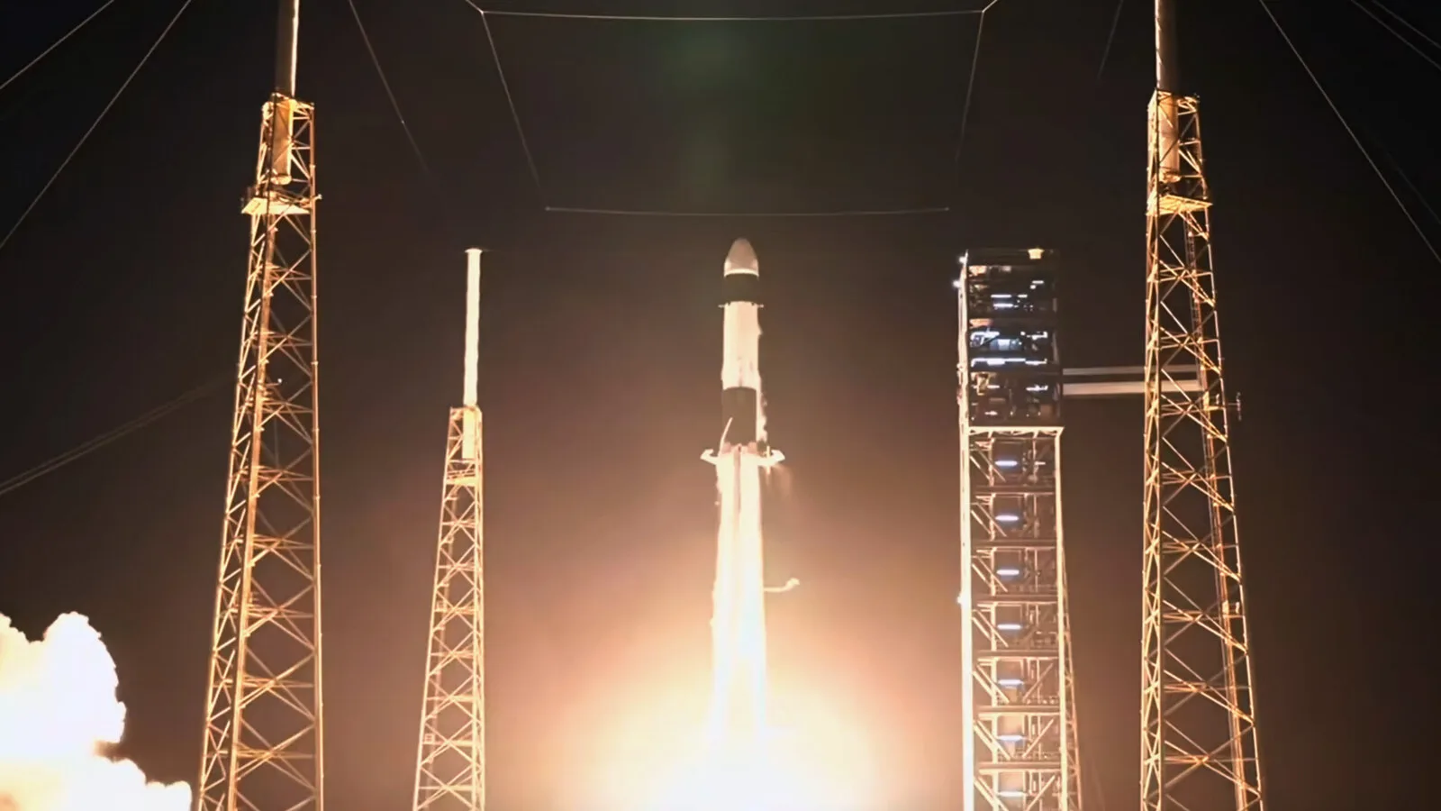 Foguete Falcon 9 a descolar de noite entre as torres de iluminação da plataforma de lançamento, com uma nuvem de fumo iluminada à esquerda