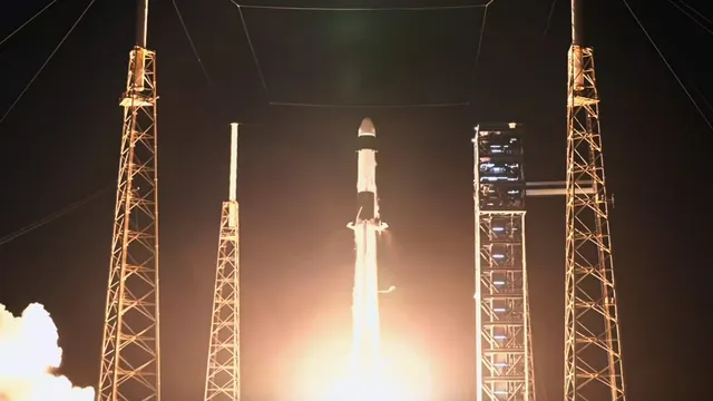 Foguete Falcon 9 a descolar de noite entre as torres de iluminação da plataforma de lançamento, com uma nuvem de fumo iluminada à esquerda
